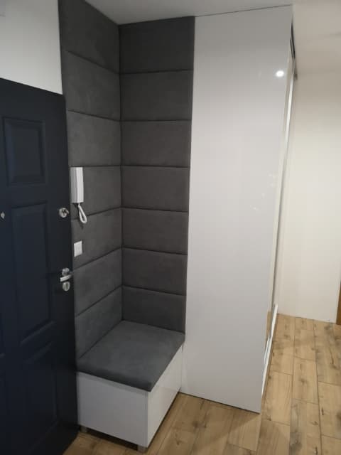 Wnęka w przedpokoju z siedziskiem i tapicerowanymi panelami ściennymi w kolorze szarym, obok szafa z białymi, błyszczącymi drzwiami.