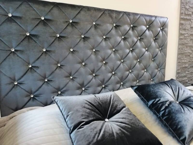 Szary, welurowy zagłówek łóżka z pikowaniem w romby i kryształkami, dopasowane poduszki dekoracyjne, fragment ściany z imitacją kamienia w tle.