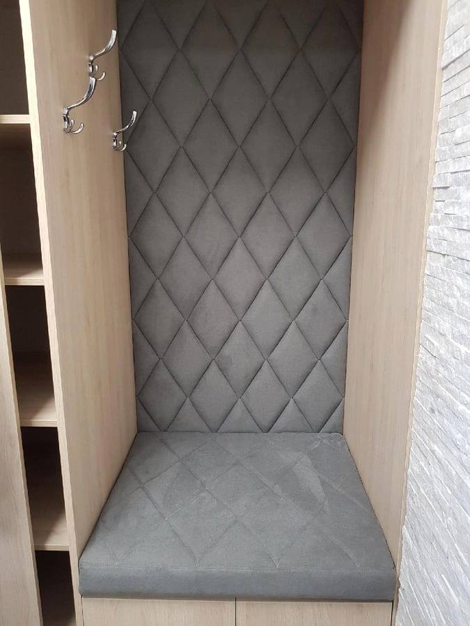 Wnęka w przedpokoju z siedziskiem i tapicerowanym panelem ściennym w szarym kolorze, pikowana w romby, z wieszakami po lewej stronie i wykończeniem z jasnego drewna.