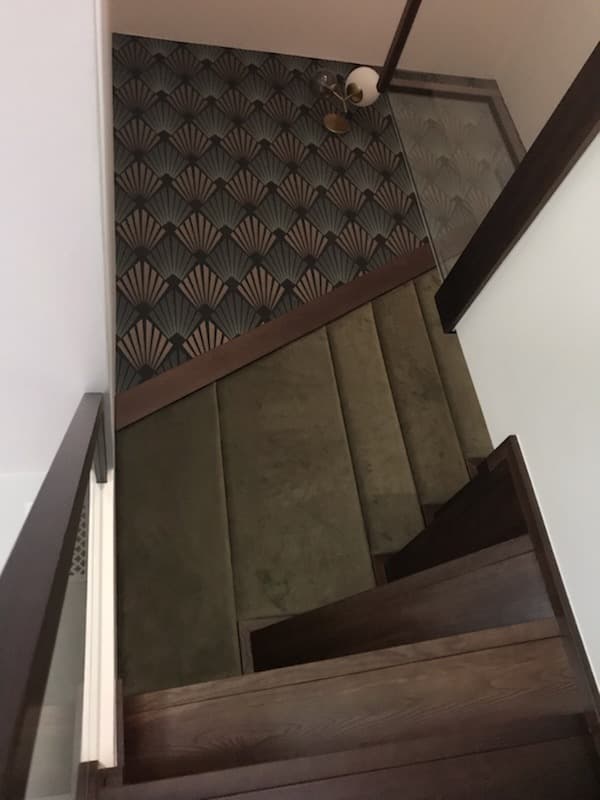 Widok z góry na drewniane schody z zielonym, tapicerowanym bieżnikiem, przylegające do ściany z wzorzystą tapetą w stylu art deco.