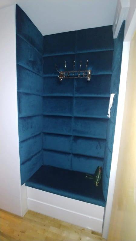 Narożne siedzisko wnękowe z tapicerowanymi panelami ściennymi w kolorze głębokiego turkusu, uzupełnione wieszakiem na ubrania w stylu retro i damską torebką w tle