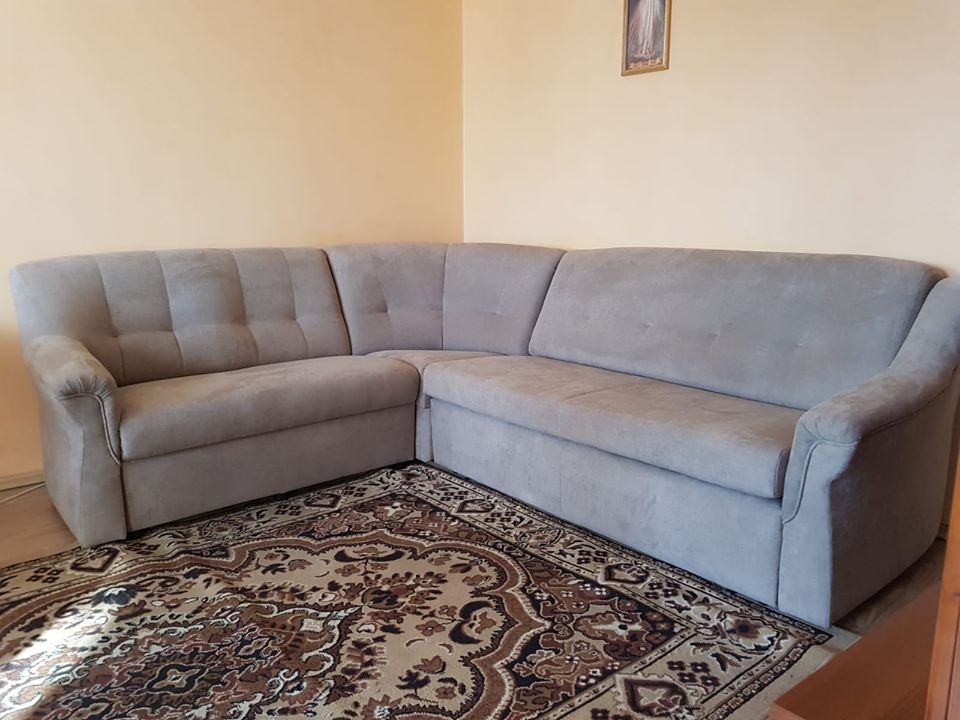 Szary narożnik z tapicerowanymi guzikami na oparciu, ustawiony na wzorzystym dywanie w beżowo-brązowych tonacjach, z obrazkiem w złotej ramce wiszącym na ścianie w tle.