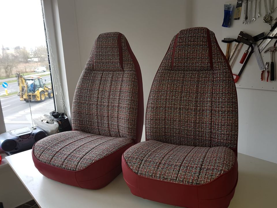 Dwa odnowione fotele samochodowe z tapicerką w drobną kratę, wykończone czerwoną skórą, widoczne na tle okna z widokiem na ulicę i narzędzi na ścianie.