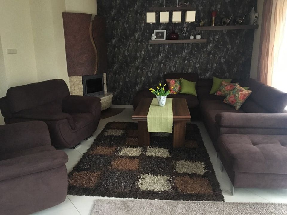 Brązowy narożnik i fotel z weluru w salonie z kominkiem, wzorzysta tapeta i dywan w odcieniach brązu, stół z żółtymi tulipanami.