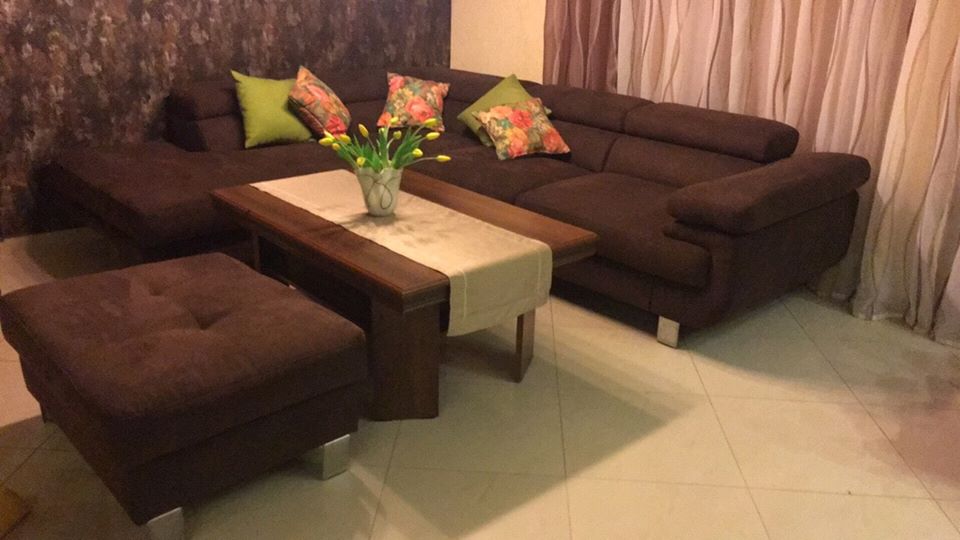 Brązowy narożnik z poduszkami w kwiaty i zielonymi, dopasowany puf z guzikiem, stolik kawowy z kremowym bieżnikiem i wazonem z żółtymi tulipanami, ustawione na jasnej podłodze.