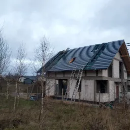 Dom w stanie surowym zamkniętym z częściowo położonym, ciemnoszarym dachem z dachówki ceramicznej, widoczne dwie drabiny opierające się o budynek, pochmurne niebo w tle.