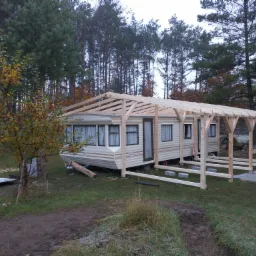 Montaż drewnianej konstrukcji zadaszenia nad przyczepą kempingową na trawiastym terenie, widoczne elementy konstrukcyjne i otaczające drzewa.