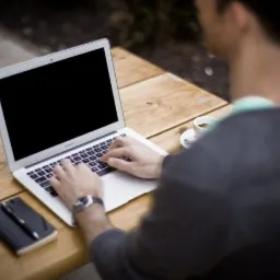 Mężczyzna pisze na laptopie na drewnianym stole, obok smartfon, notatnik z długopisem i filiżanka kawy.