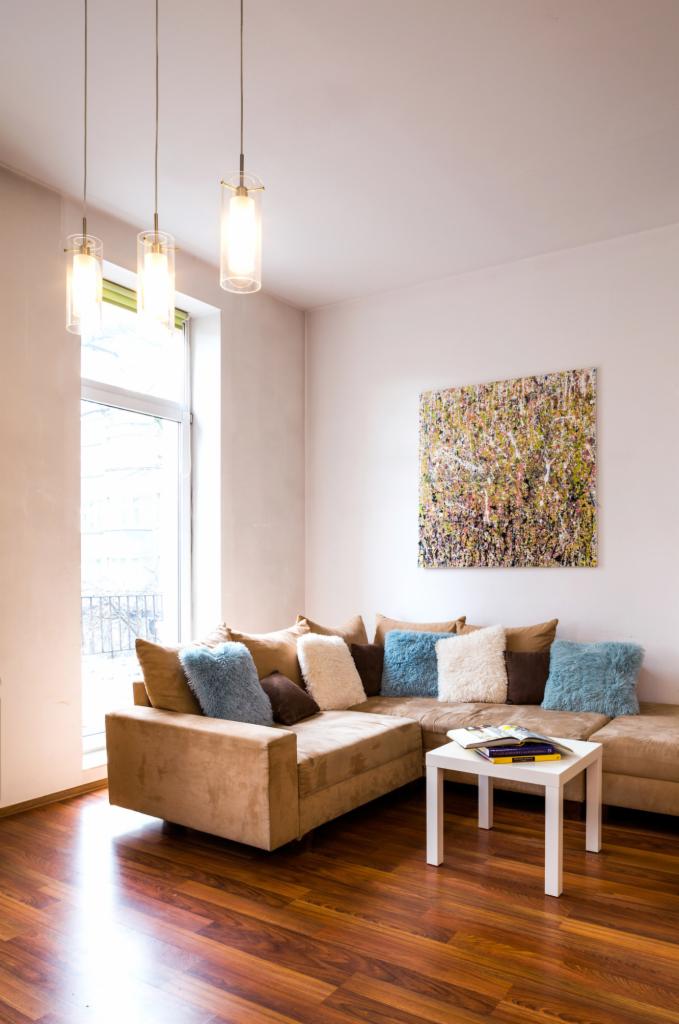 Jasne wnętrze pokoju dziennego z narożną kanapą, dekoracyjnymi poduszkami, obrazem na ścianie i trzema wiszącymi lampami o cylindrycznym kształcie.