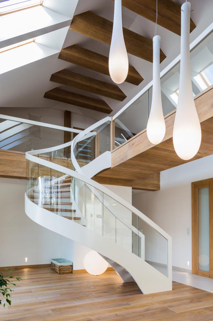 Nowoczesne wnętrze z krętymi białymi schodami z przeszkloną balustradą, drewnianym sufitem z belkami i trzema podłużnymi lampami wiszącymi.