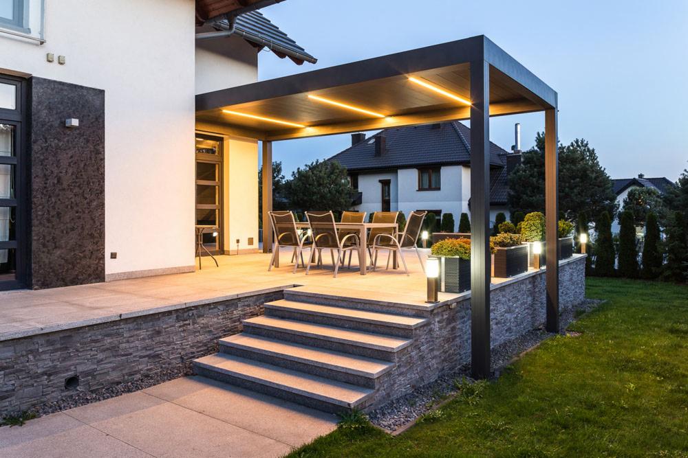 Zadaszony taras z oświetleniem LED, meblami ogrodowymi i schodami, widok na dom w tle.