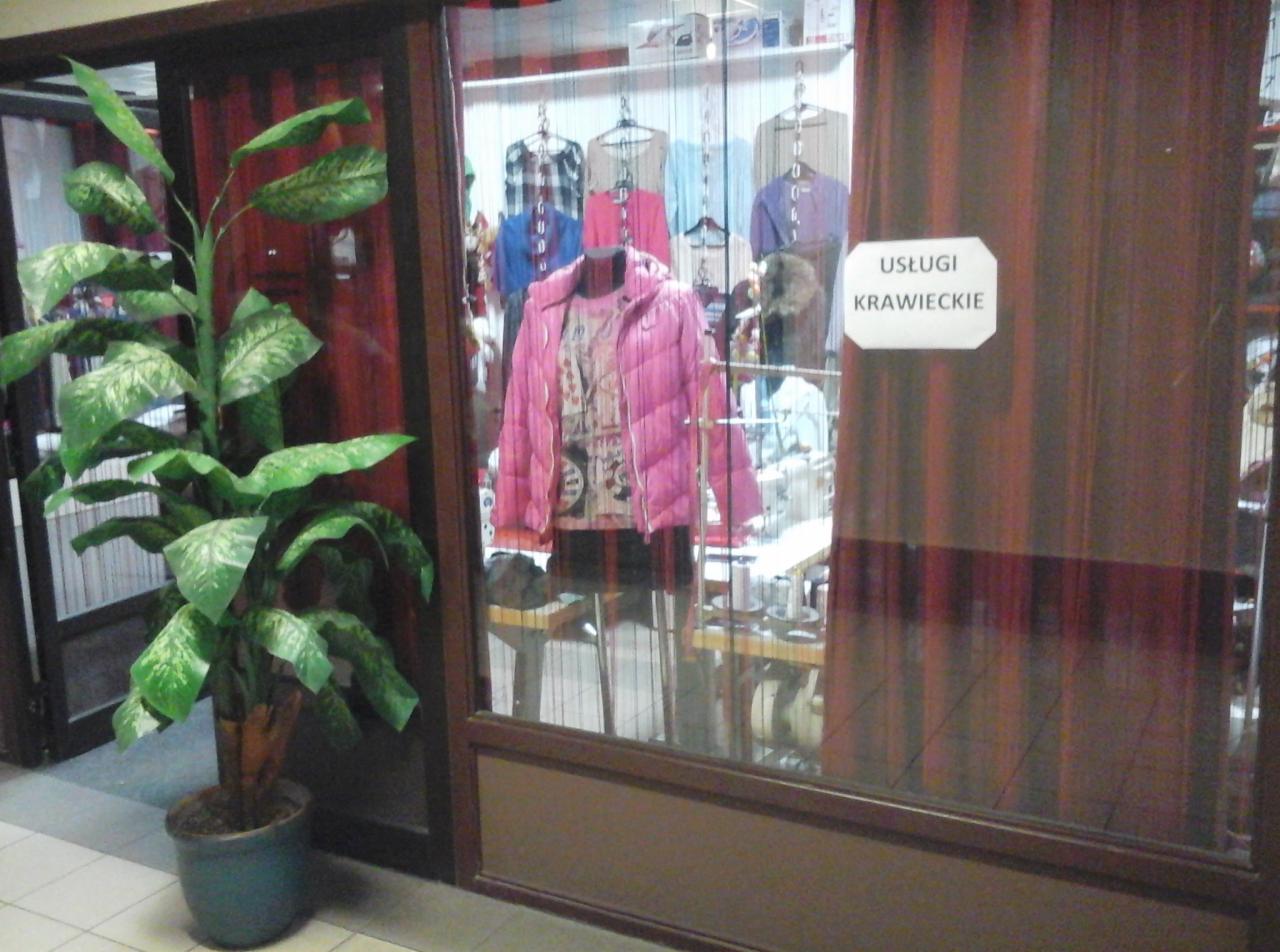 Wystawa zakładu z różową kurtką na manekinie, w tle wiszące ubrania i maszyny do szycia, z boku doniczka z dużą rośliną.