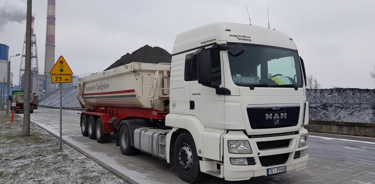 Ciężarówka MAN z naczepą wypełnioną ziemią lub węglem, zaparkowana na tle kominów przemysłowych i hałdy, widoczny znak drogowy.