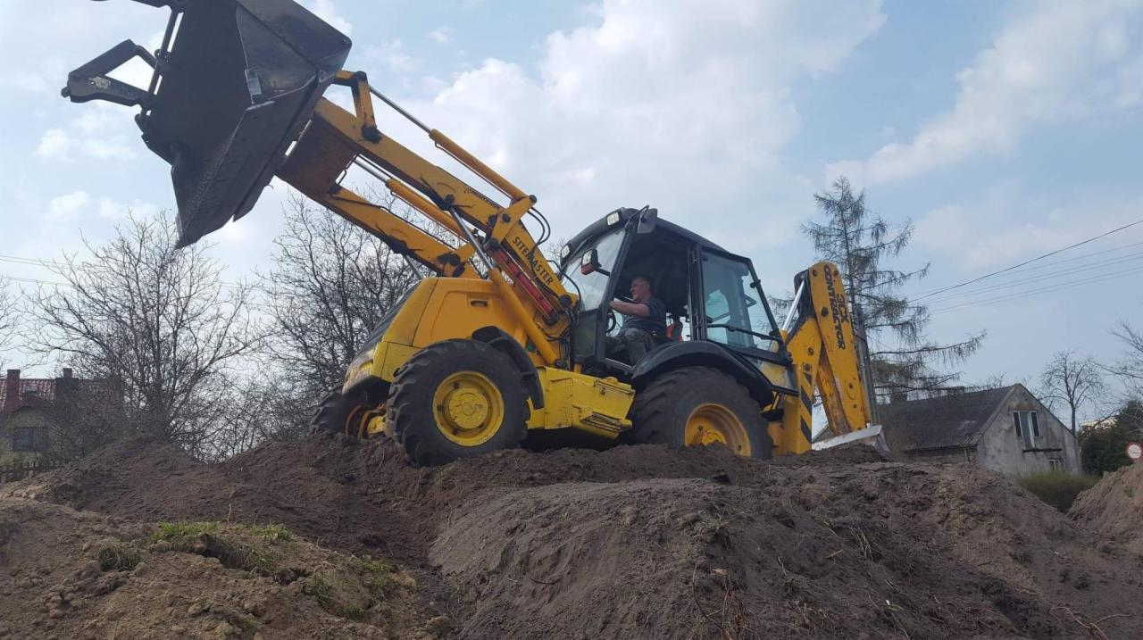 Żółta koparko-ładowarka na wzniesieniu z ziemi w Chmielniku, z widocznym operatorem w kabinie i podniesioną łyżką z przodu, w tle zabudowania mieszkalne.