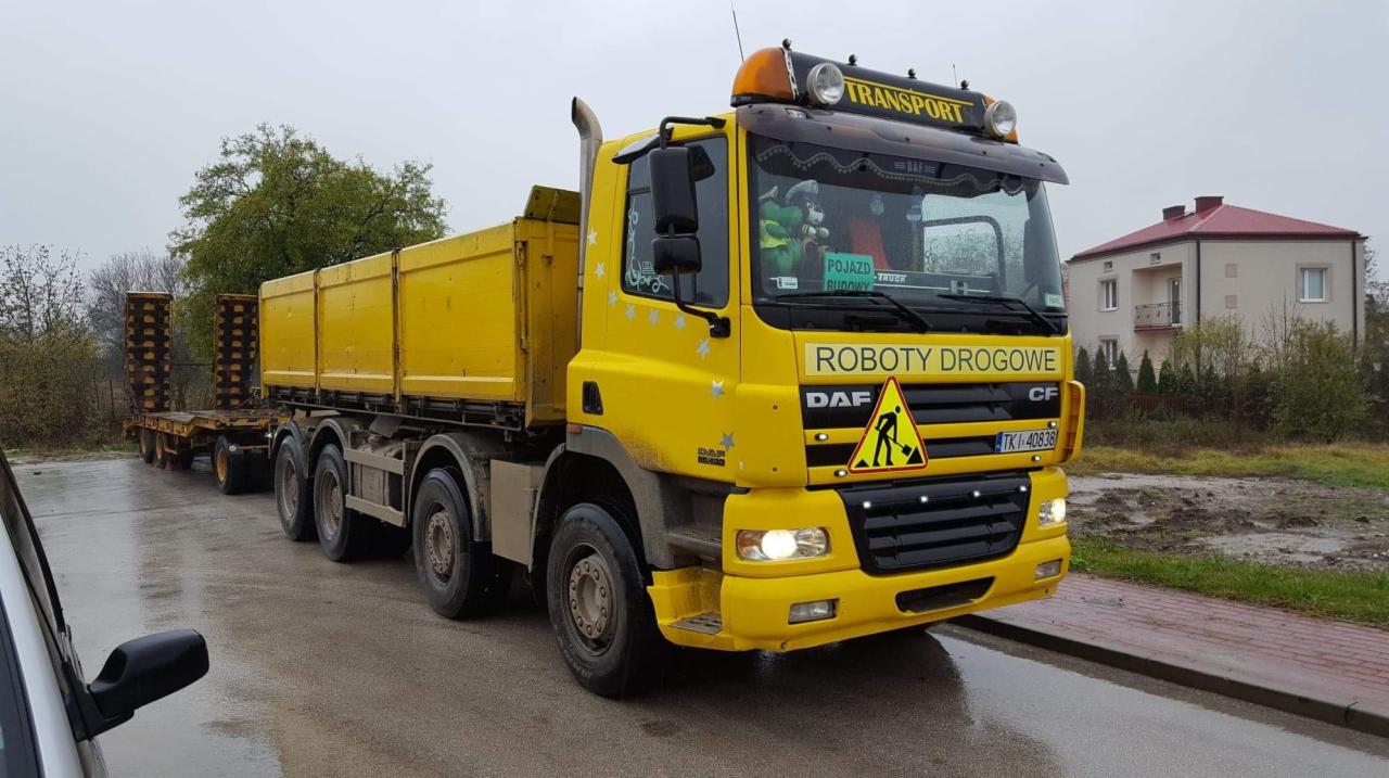 Żółta ciężarówka DAF z naczepą niskopodwoziową i napisem 'ROBOTY DROGOWE' na tle szarego nieba i domu.