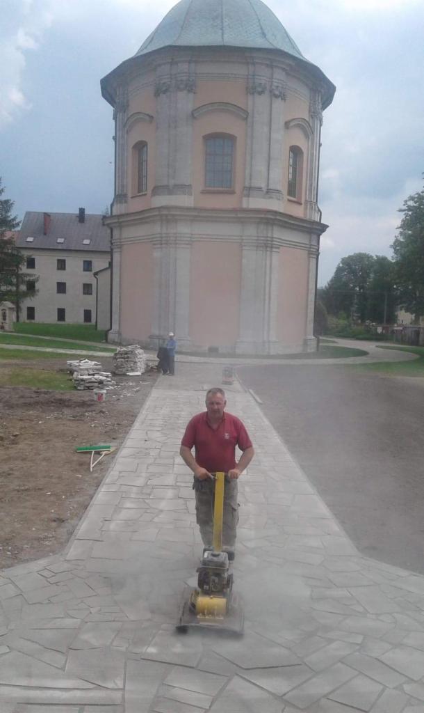 Pracownik zagęszcza wibropraską ułożoną ścieżkę z nieregularnych płyt kamiennych, w tle budynek sakralny z kopułą.
