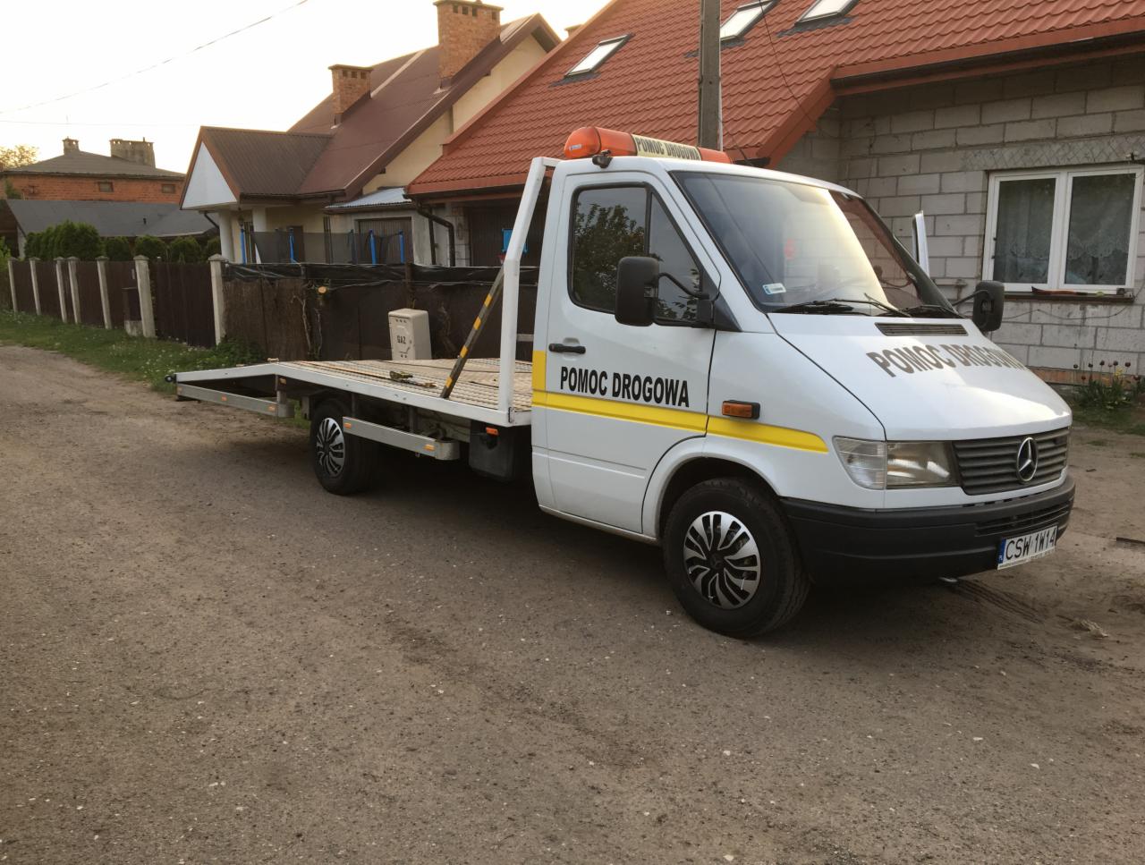 Biała laweta Mercedes Sprinter z napisem 'POMOC DROGOWA' na tle domów jednorodzinnych i polnej drogi.