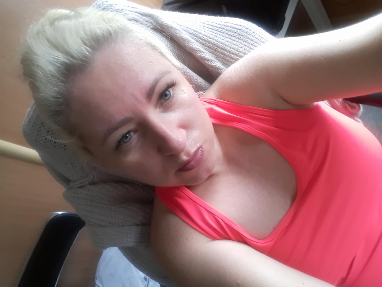 Portret kobiety w różowej koszulce, leżącej na krześle z szarą narzutą, z bląd włosami i niebieskimi oczami.