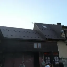 Dom z nowym, brązowym dachem blaszanym z oknem dachowym, antena satelitarna i komin, widoczne osoby przy pracy.