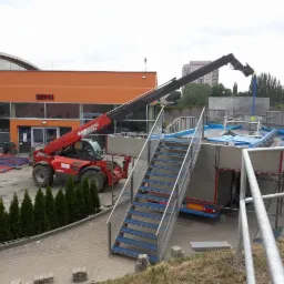 Czerwony podnośnik teleskopowy Maniscopic umieszcza element konstrukcyjny na platformie z niebieskimi akcentami, obok schody z niebieskimi stopniami, w tle budynek z zegarem.