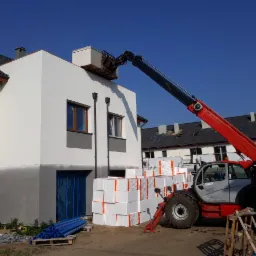 Czerwony podnośnik teleskopowy umieszcza paletę z materiałem izolacyjnym na dachu domu w trakcie budowy, z zapasem styropianu u podstawy i niebieskimi rurami na palecie z boku.