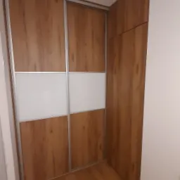 Wnętrze z zabudowaną szafą przesuwną z drewnopodobnymi frontami i białym pasem, dopasowaną do skosu pomieszczenia.