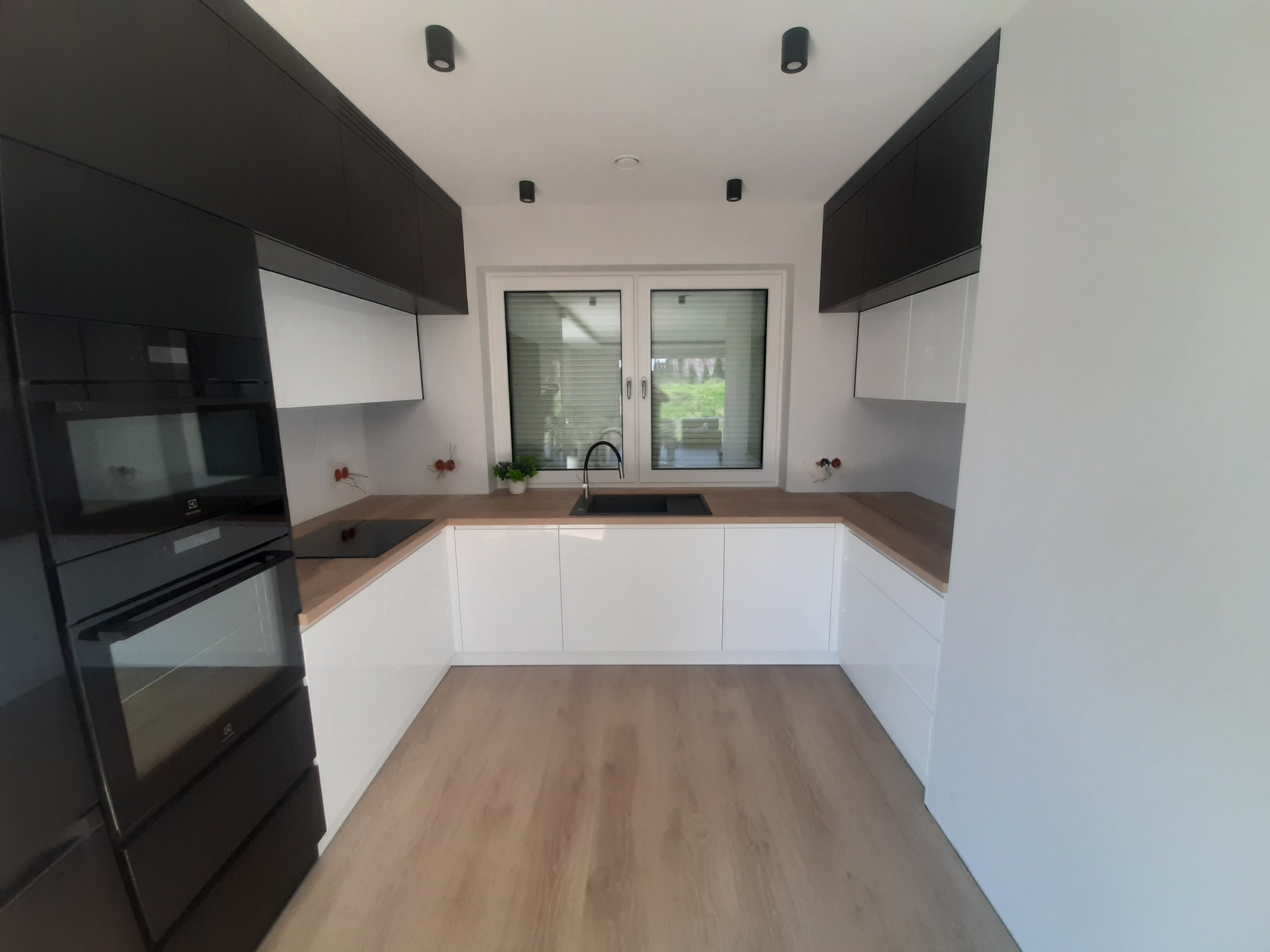 Nowoczesna kuchnia w kształcie litery U z białymi frontami i drewnianym blatem, czarne szafki wiszące, oświetlenie punktowe, okno z żaluzjami i drewniana podłoga.