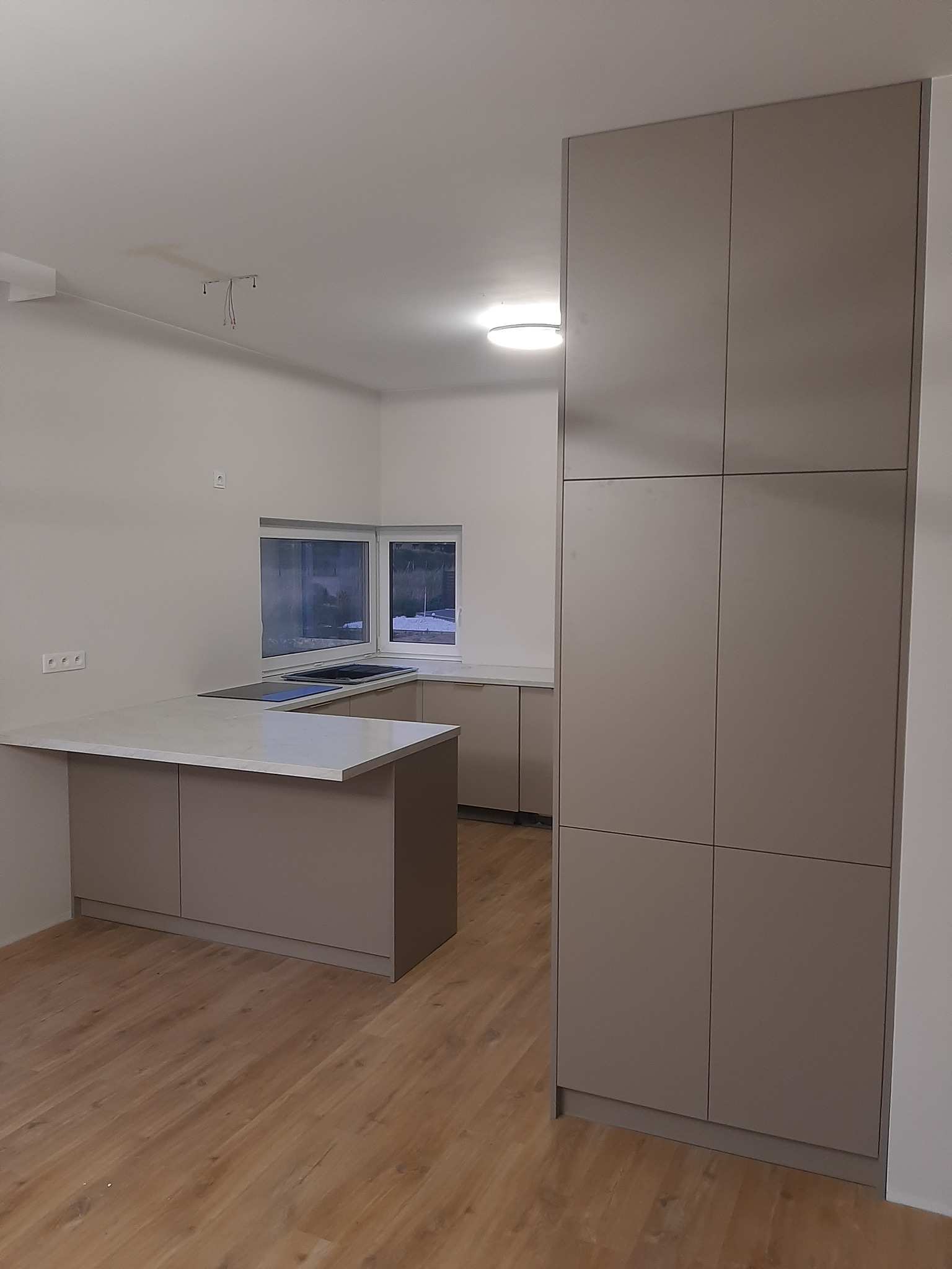 Nowoczesna kuchnia z wyspą i wysoką zabudową w odcieniach beżu, z drewnianą podłogą i oknem narożnym. Minimalistyczny design, brak uchwytów.