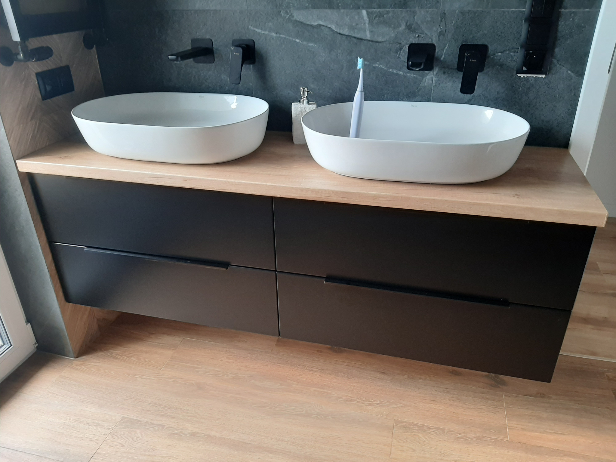 Nowoczesna łazienka z dwiema owalnymi umywalkami nablatowymi, czarną szafką z długimi uchwytami i drewnianym blatem. Na blacie mydło w płynie i szczoteczka do zębów.