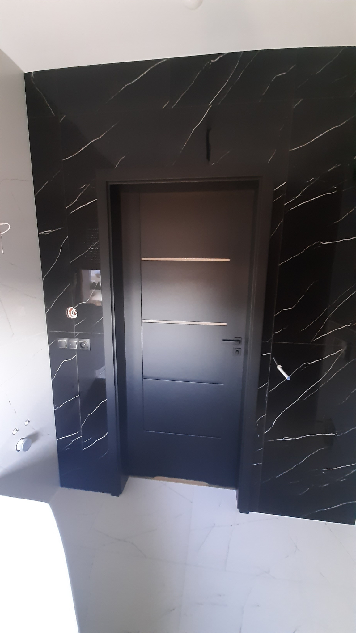Ciemne drzwi wewnętrzne z trzema poziomymi frezami, osadzone w ścianie wyłożonej czarnym marmurem z białymi żyłami. Minimalistyczny design, nowoczesne wykończenie.