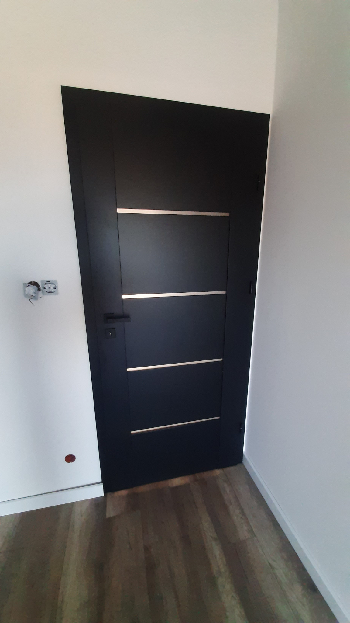 Czarne drzwi wewnętrzne z trzema poziomymi, jasnymi paskami, zamontowane w białej ścianie. Widoczna klamka i fragment drewnianej podłogi.