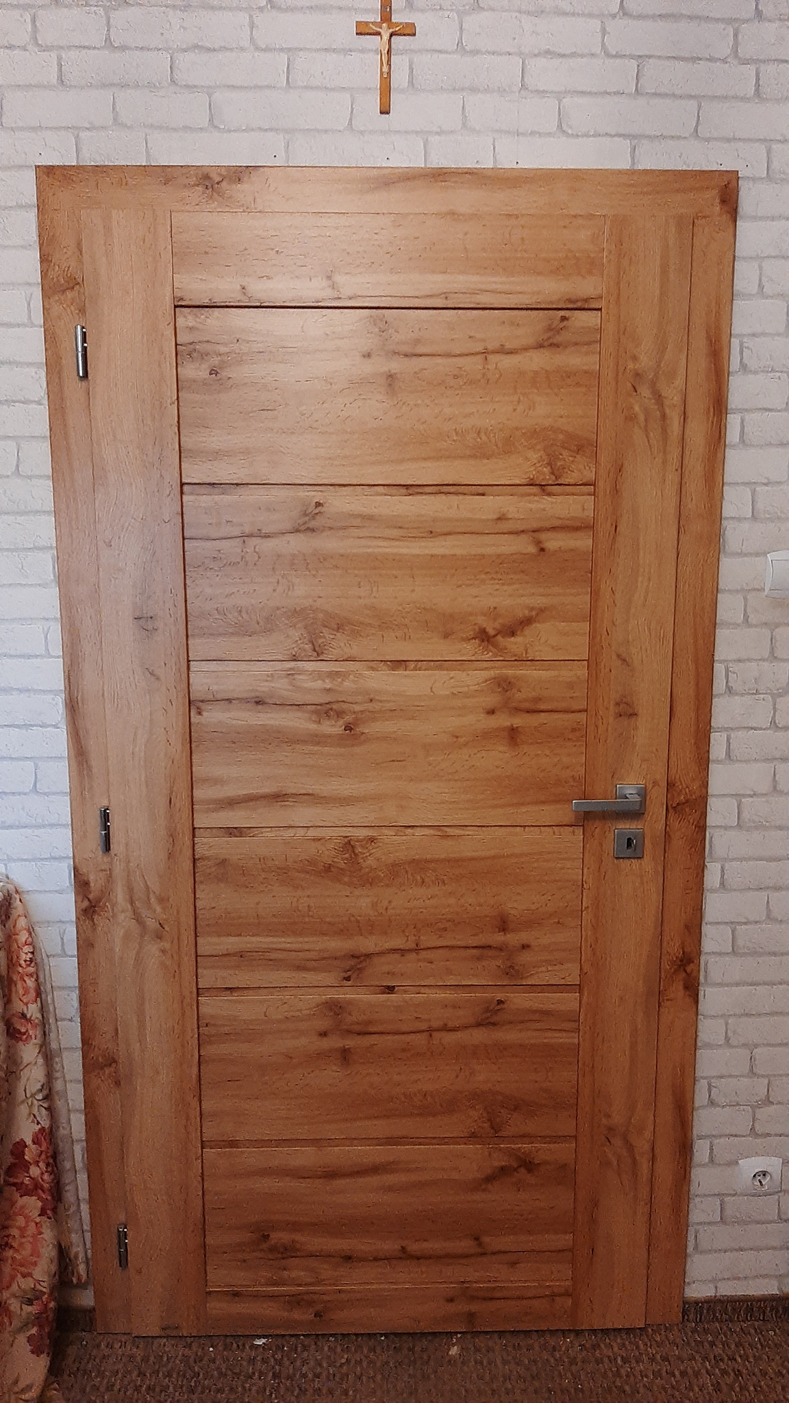 Drewniane drzwi wewnętrzne z poziomymi panelami, widoczne na tle białej ceglanej ściany z krzyżem powyżej. Klamka po prawej stronie.