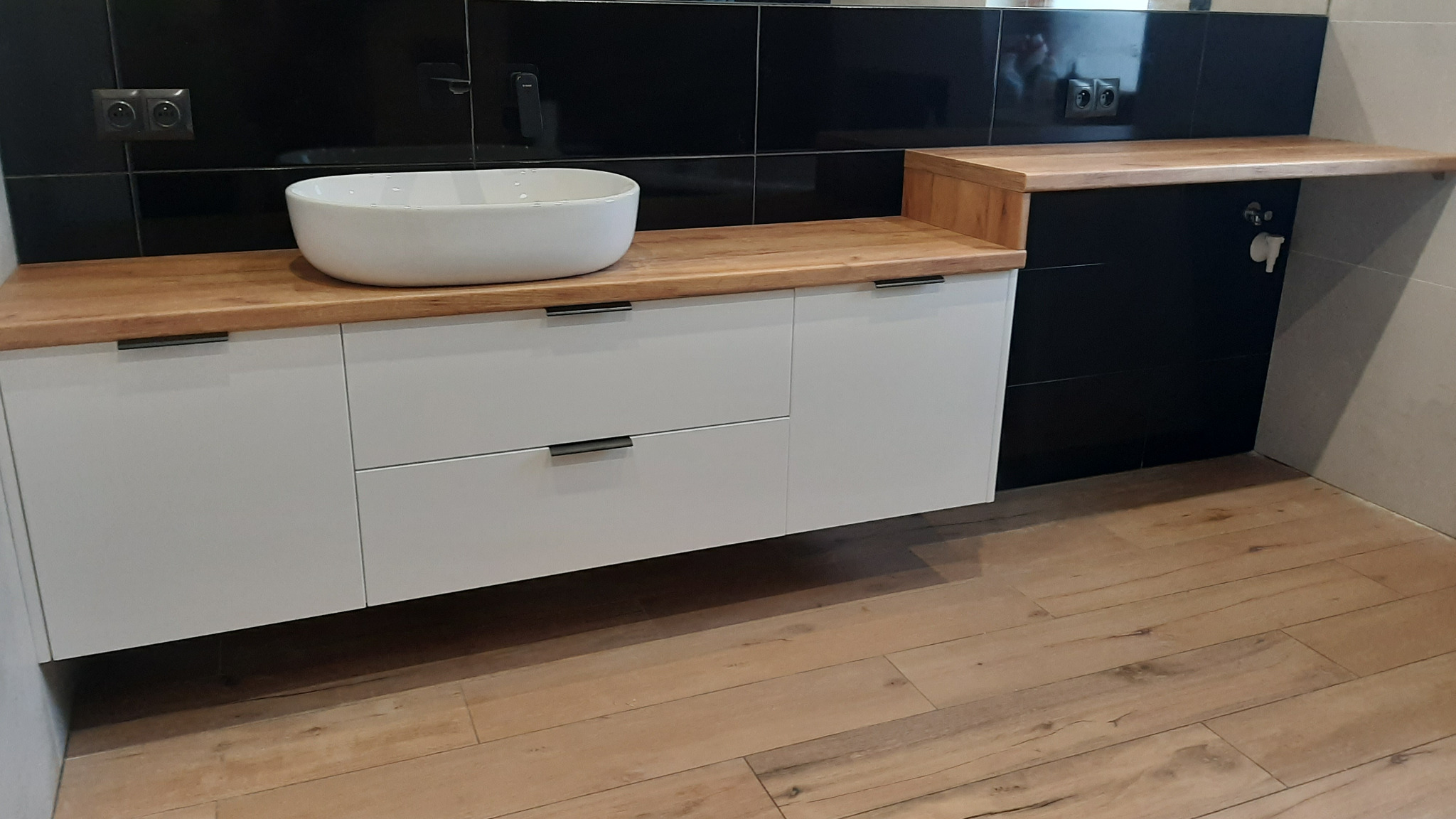 Biała szafka łazienkowa z drewnianym blatem i umywalką nablatową na tle czarnych płytek. Ujęcie z perspektywy podłogi, ukazujące detale wykończenia.