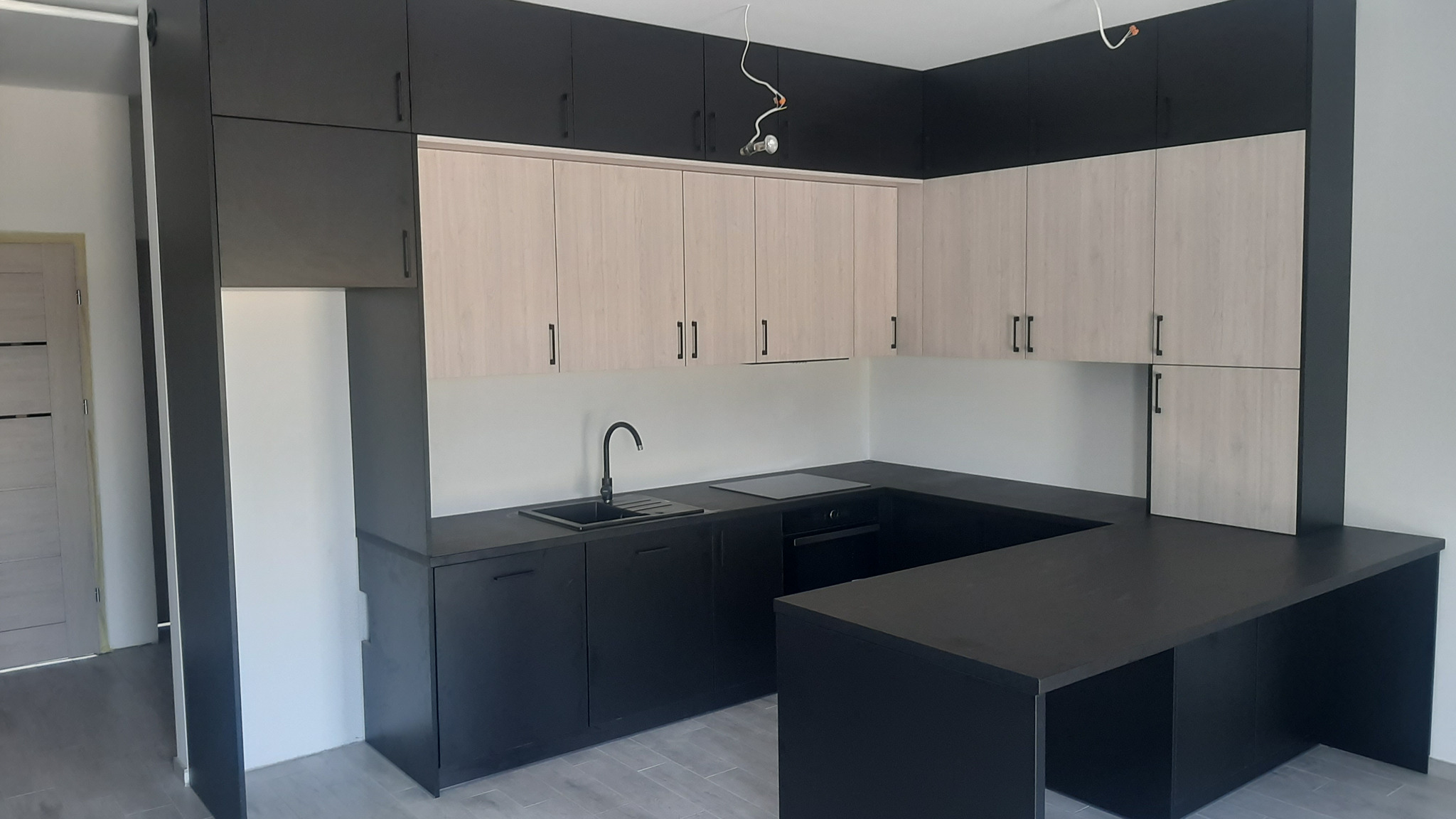 Nowoczesna kuchnia w odcieniach czerni i beżu, z minimalistycznymi frontami szafek i blatem. Widoczne oświetlenie sufitowe i fragment drzwi w tle.