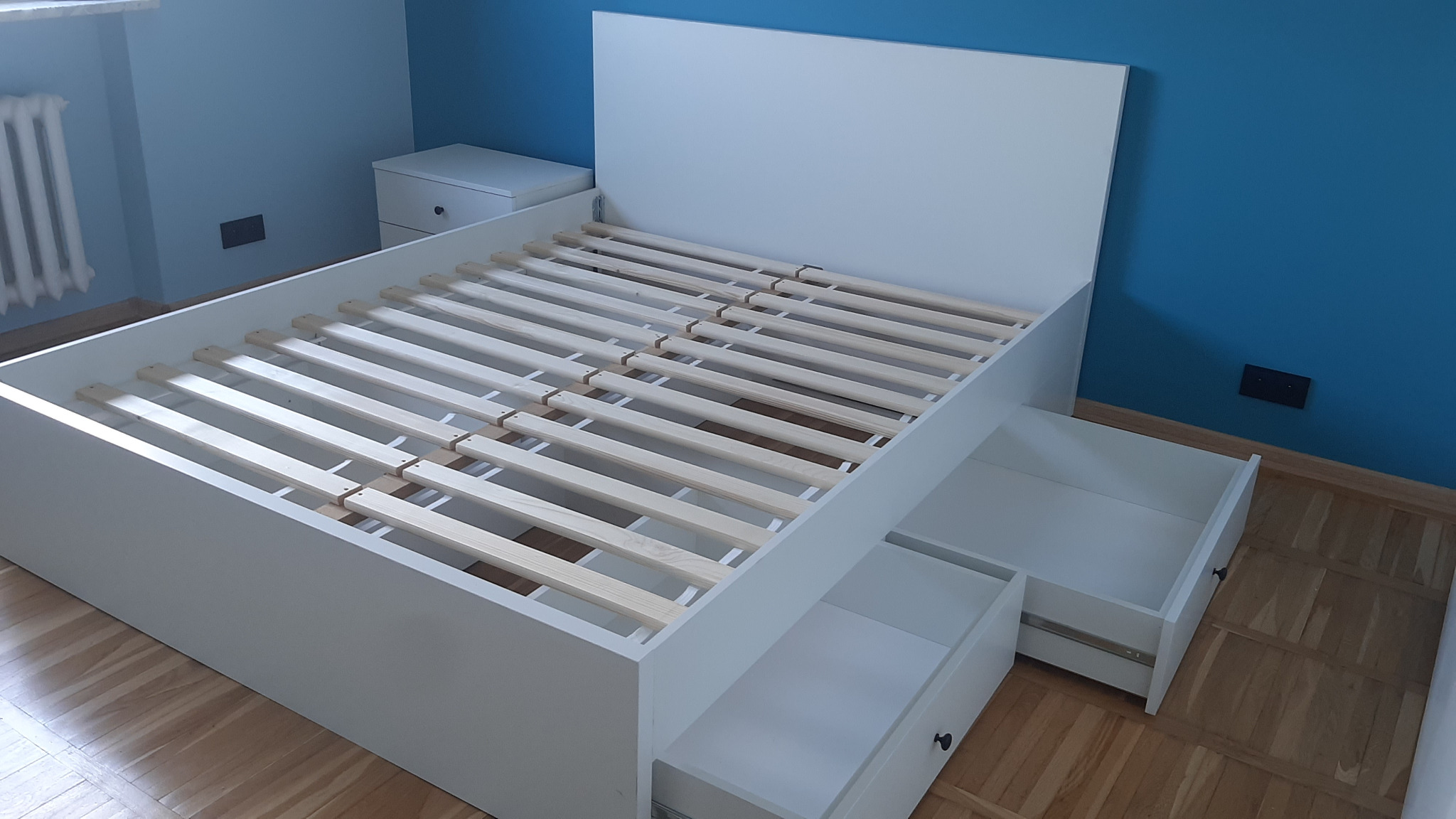 Białe łóżko z drewnianym stelażem i wysuwanymi szufladami, ustawione na parkiecie w pokoju z niebieskimi ścianami. Widoczny fragment grzejnika i szafki nocnej.