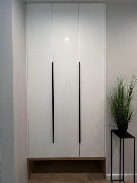 Biała, wysoka szafa z połyskiem i długimi, czarnymi uchwytami, umieszczona na drewnianej podstawie z otwartą przestrzenią. Obok stoi czarny kwietnik z zieloną rośliną.