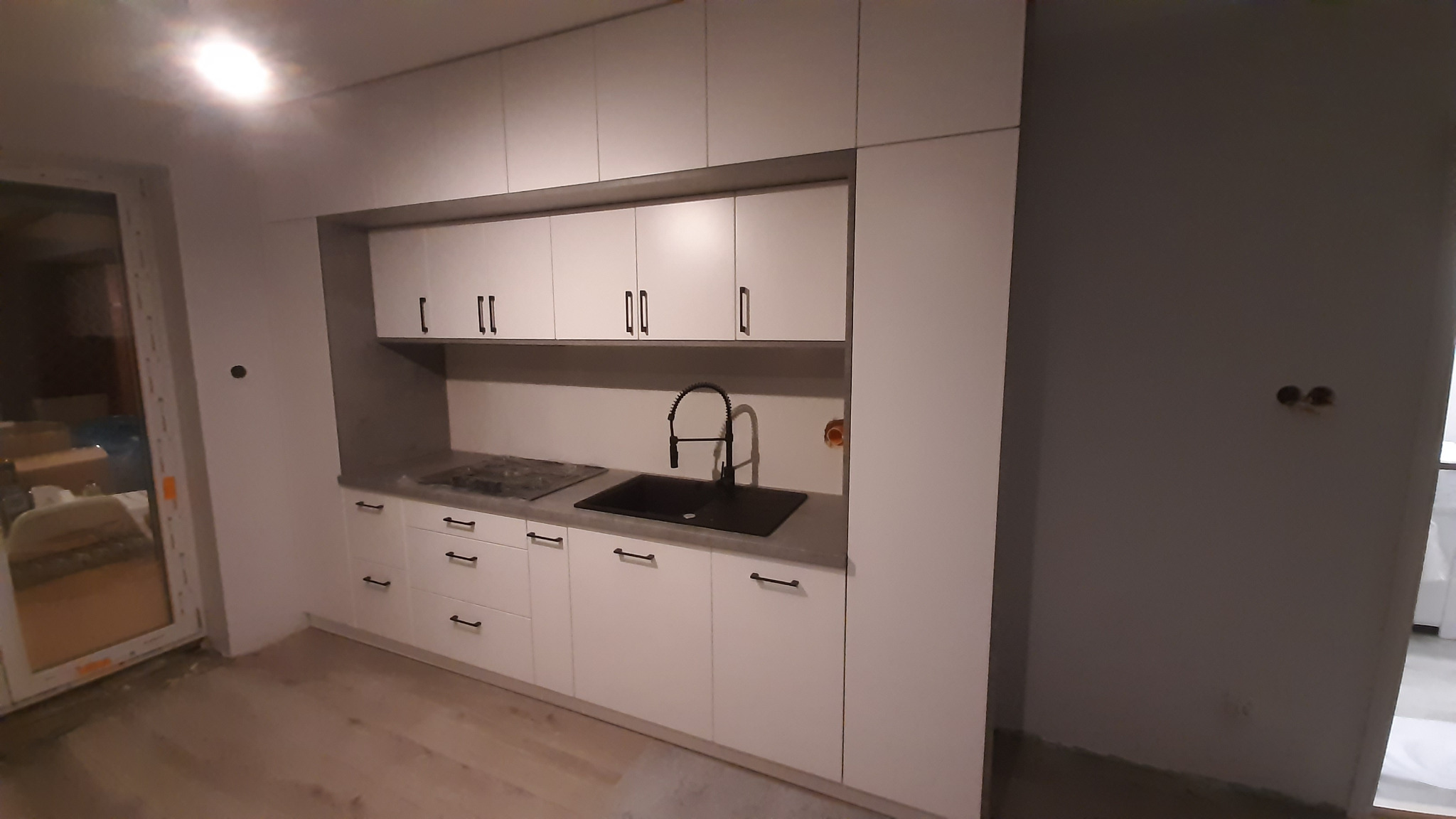 Nowoczesna, biała zabudowa kuchenna z czarnymi akcentami: minimalistyczne uchwyty, bateria i zlew. Szara ściana nad blatem. Widok na wnętrze w trakcie wykańczania.