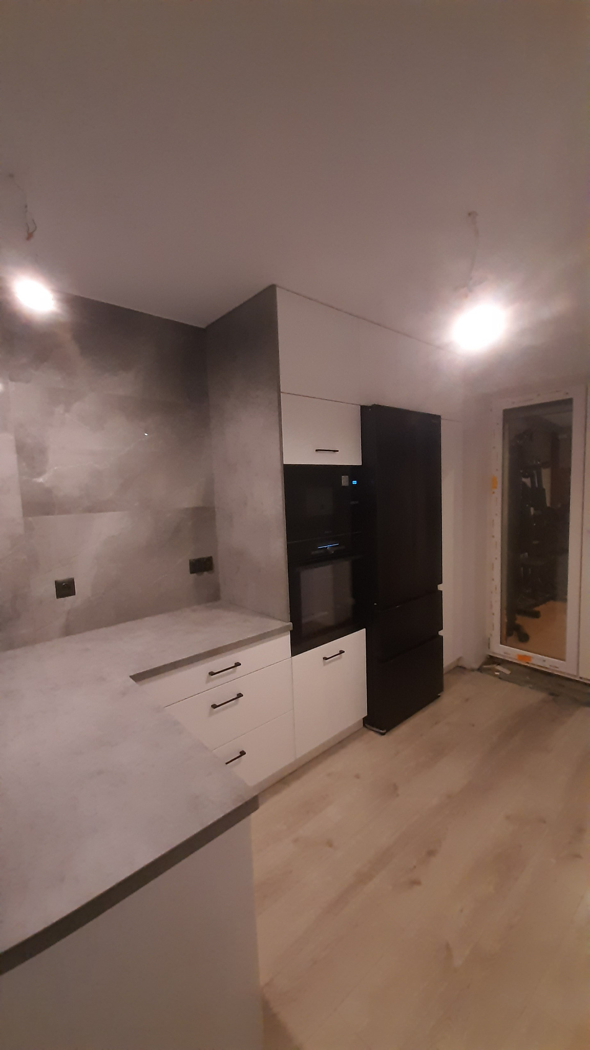 Nowoczesna kuchnia z białymi szafkami, szarym blatem i czarną lodówką w zabudowie. Minimalistyczny design, jasne drewniane podłogi, okno w tle.