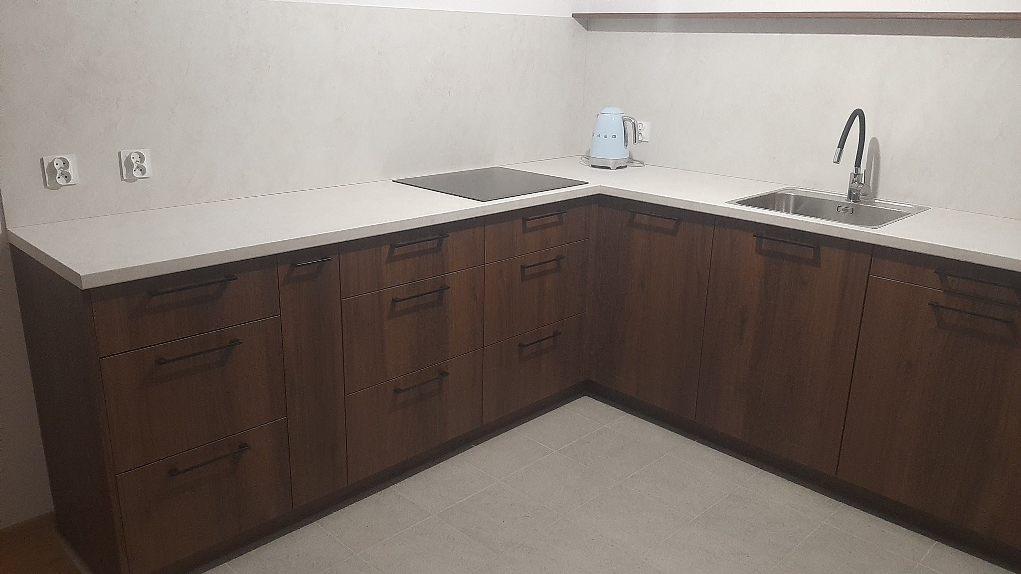 Kątowe ujęcie brązowej zabudowy kuchennej z minimalistycznymi uchwytami, jasnym blatem i płytą indukcyjną. W tle widoczne gniazdka elektryczne i czajnik.