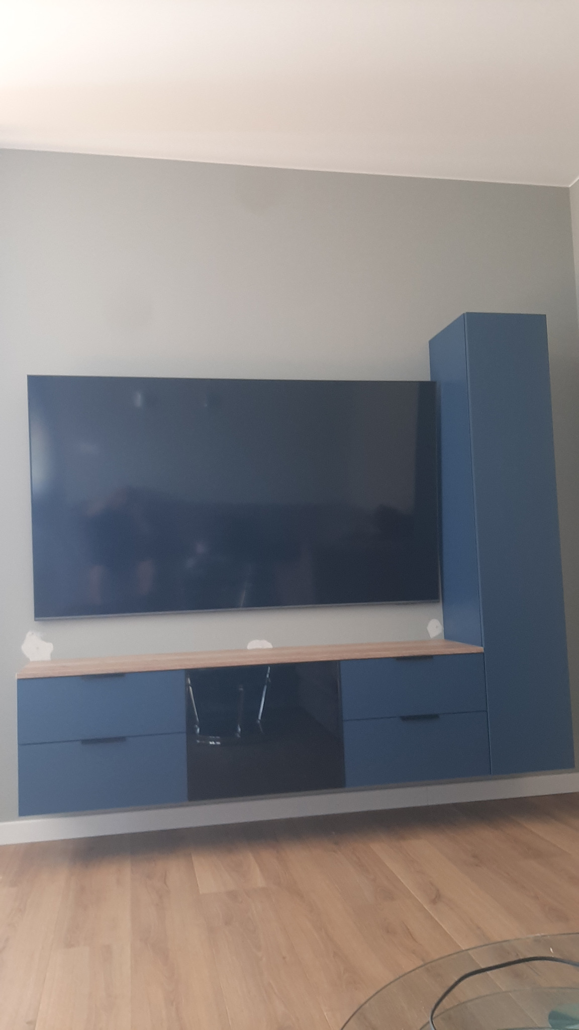 Niebieska szafka RTV z szufladami i szafką stojącą, z drewnianym blatem, zamontowana na szarej ścianie, widok z dołu, na podłodze panele.