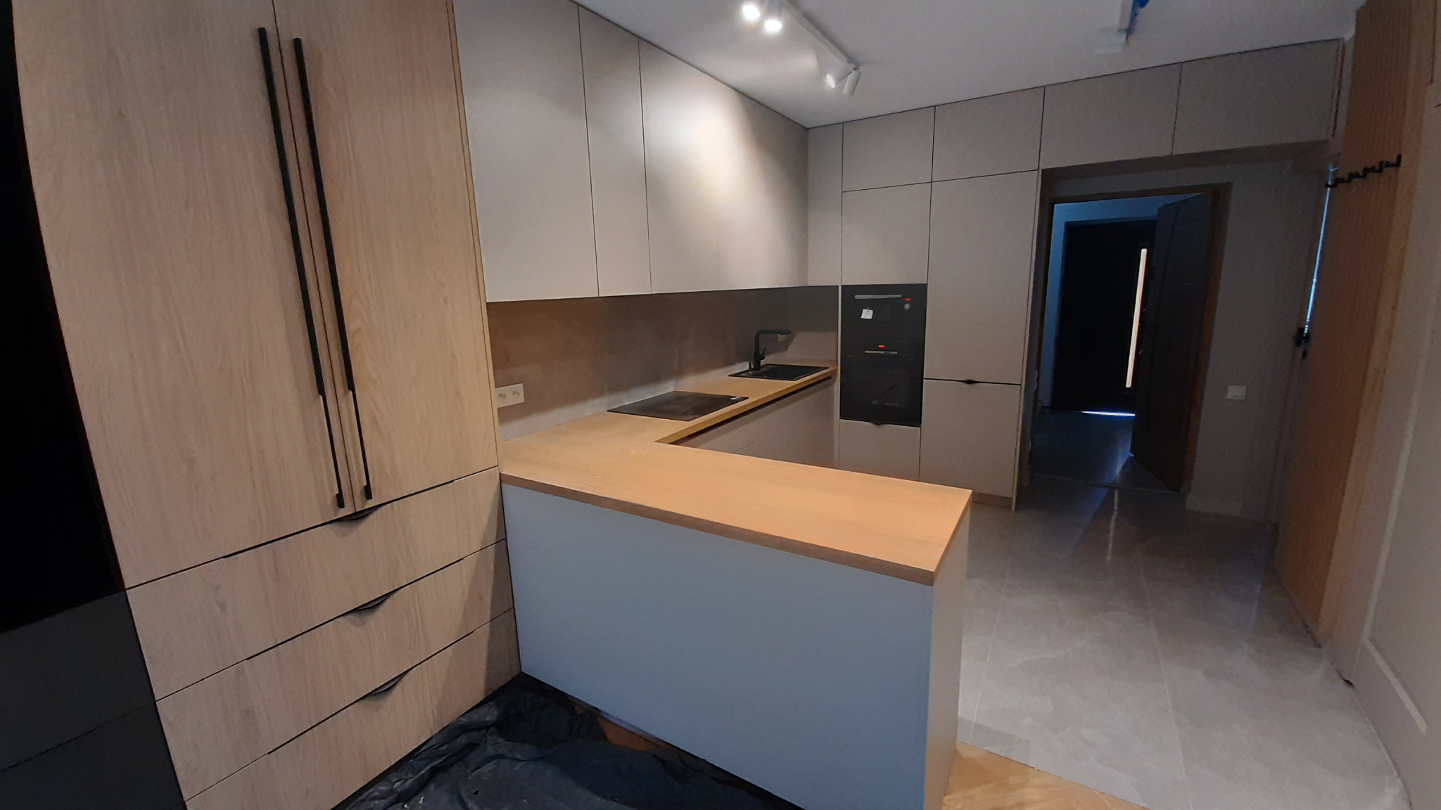 Nowoczesna kuchnia z drewnianym blatem i szarymi frontami szafek, czarne, długie uchwyty, oświetlenie punktowe, widok na otwarte drzwi do innego pomieszczenia.