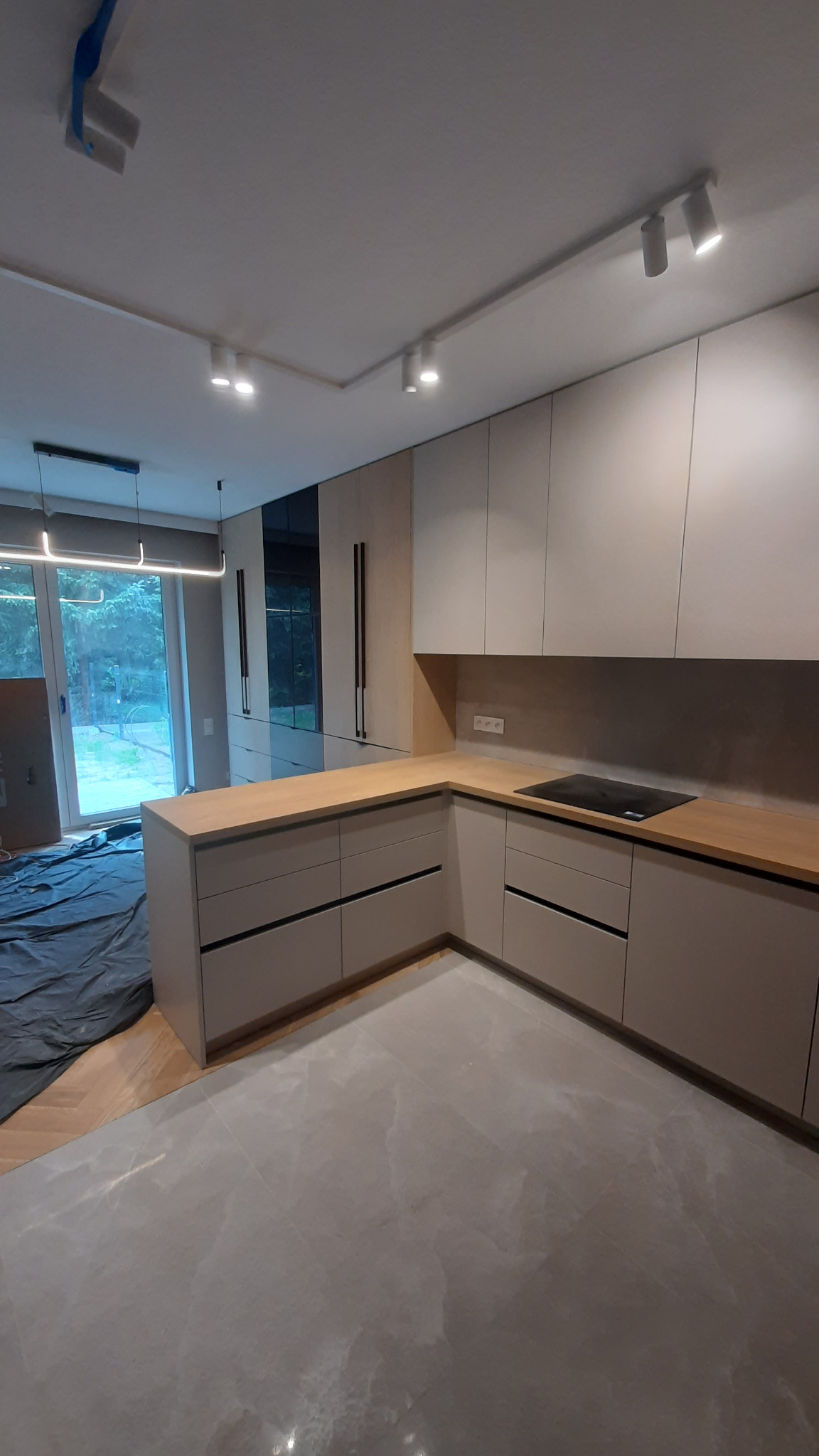 Nowoczesna kuchnia w odcieniach beżu i szarości z drewnianym blatem, oświetleniem punktowym i dużym oknem balkonowym w tle. Minimalistyczny design.