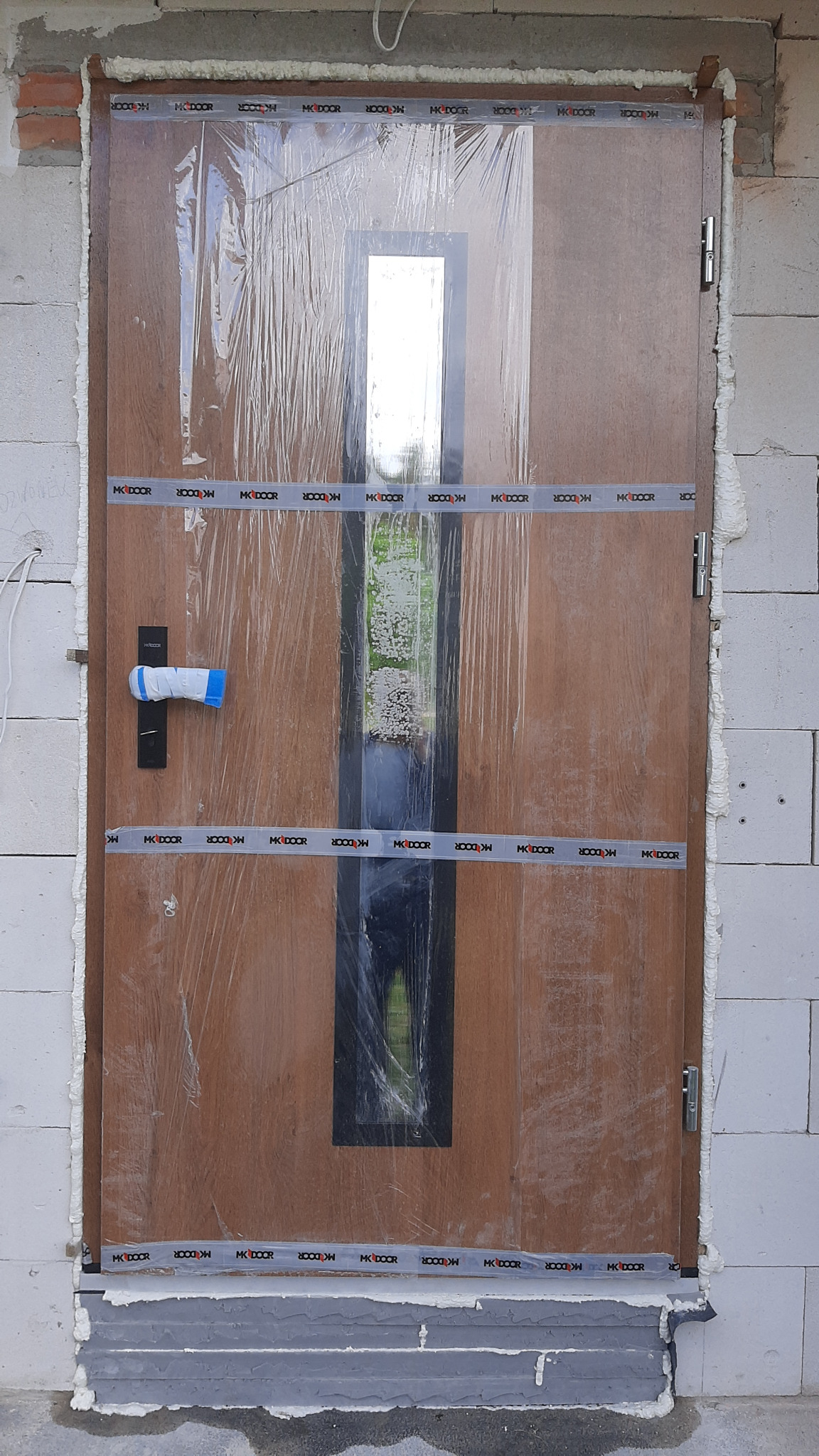 Brązowe drzwi zewnętrzne z szybą, zabezpieczone folią i taśmą montażową z logo, osadzone w murze z białych bloczków. Widoczna piana montażowa i szara podstawa.