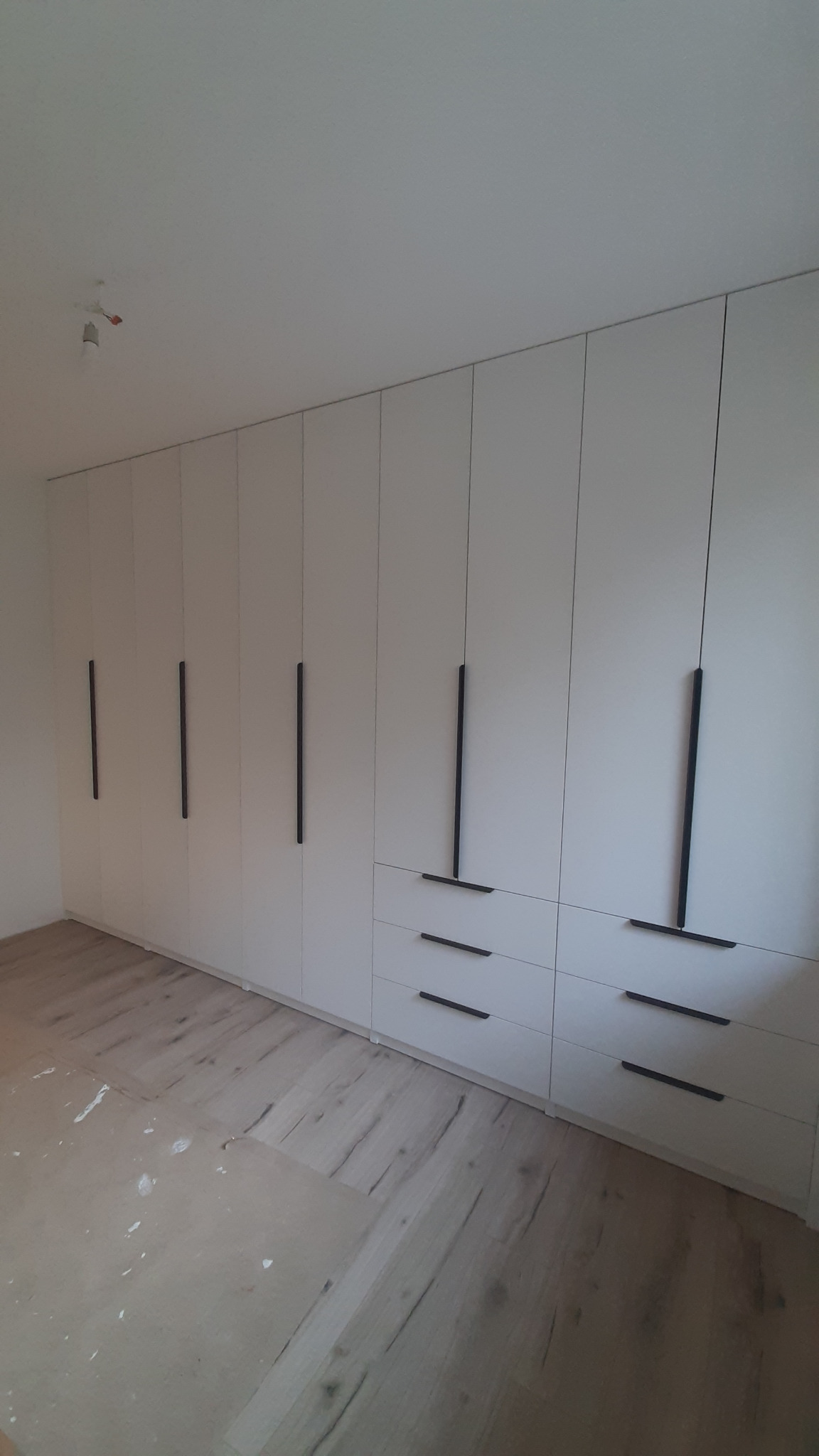 Biała szafa wnękowa z długimi, czarnymi uchwytami, wkomponowana w jasne wnętrze. Widoczne panele podłogowe i sufit z wystającą instalacją elektryczną.