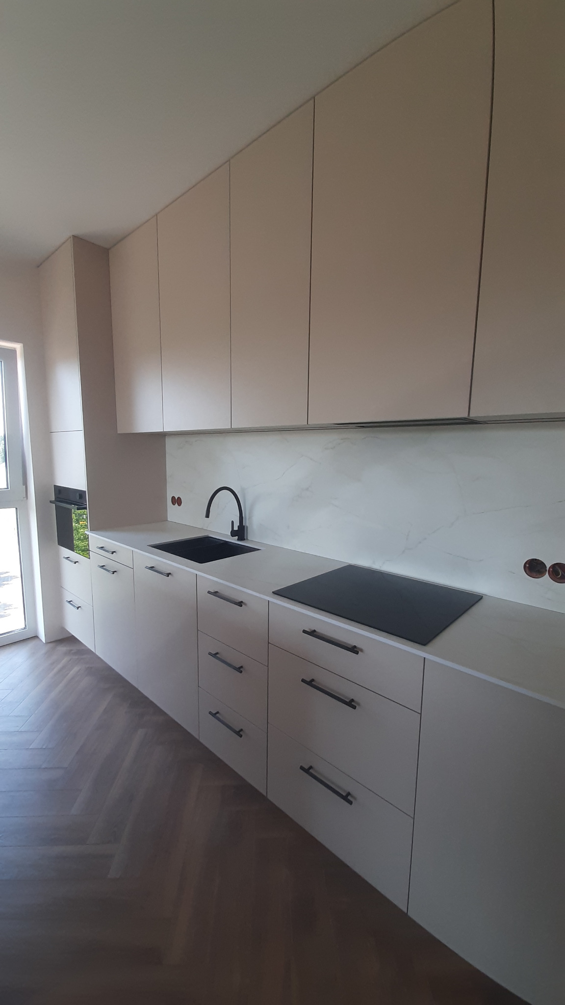 Jasna kuchnia z czarnymi akcentami: minimalistyczne szafki, czarny zlew i płyta indukcyjna na blacie z marmurowym panelem. Parkiet w jodełkę na podłodze.