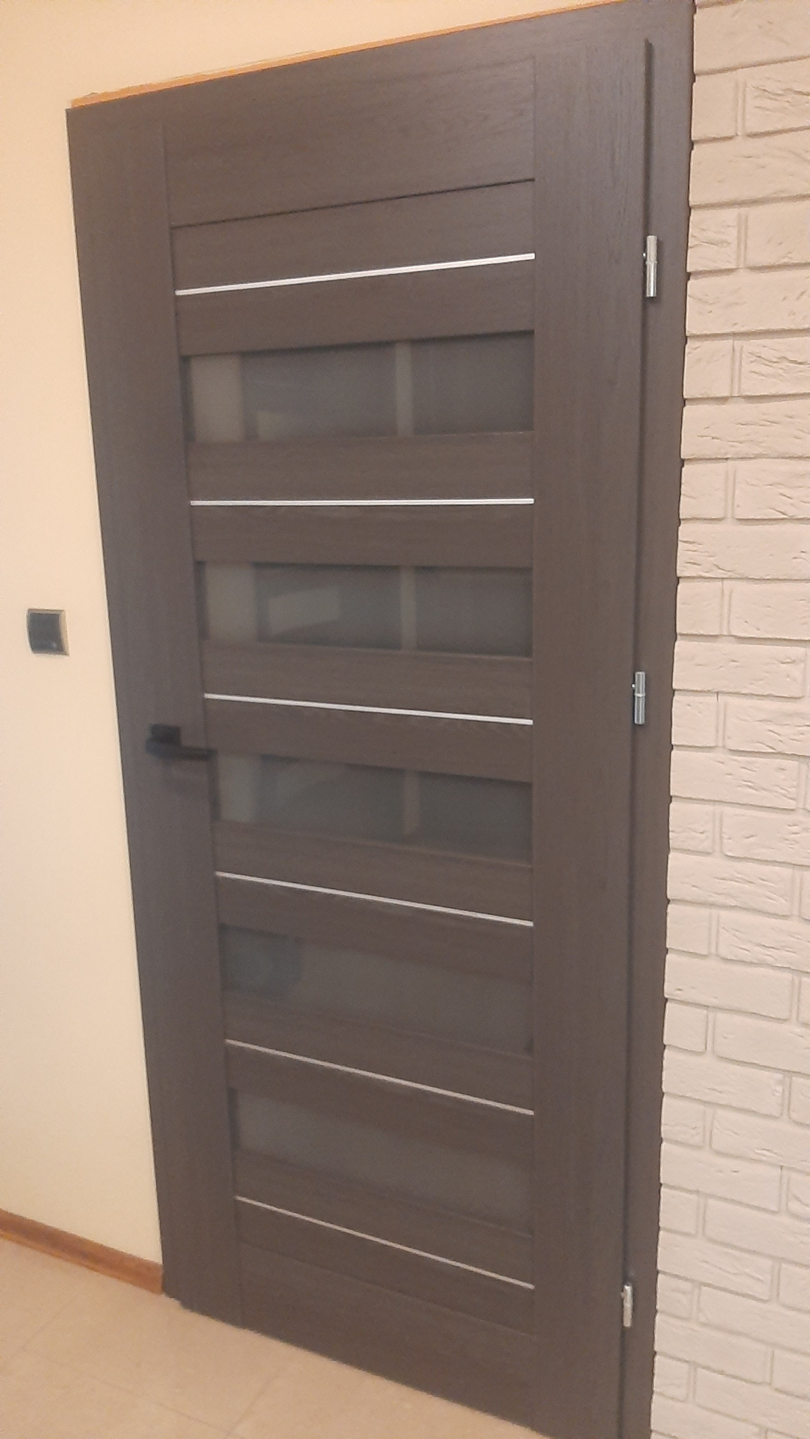 Ciemne drzwi wewnętrzne z przeszkleniami, z klamką po lewej stronie, zamontowane w Nowym Tomyślu. Obok ściana z białej cegły.