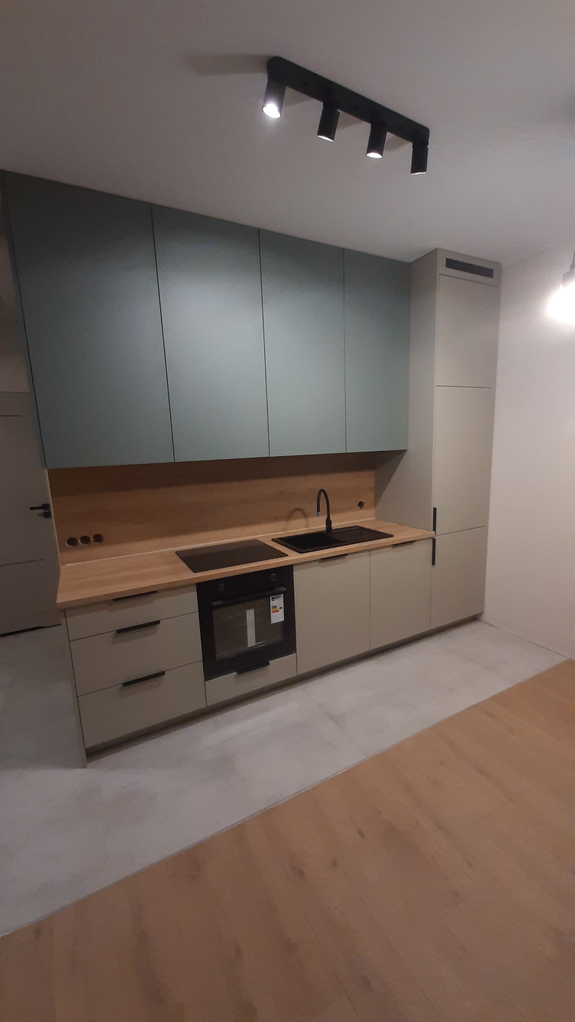 Nowoczesna kuchnia z górnymi szafkami w odcieniach zieleni i dolnymi w beżu, drewniany blat, czarny zlew i bateria, oświetlenie punktowe.