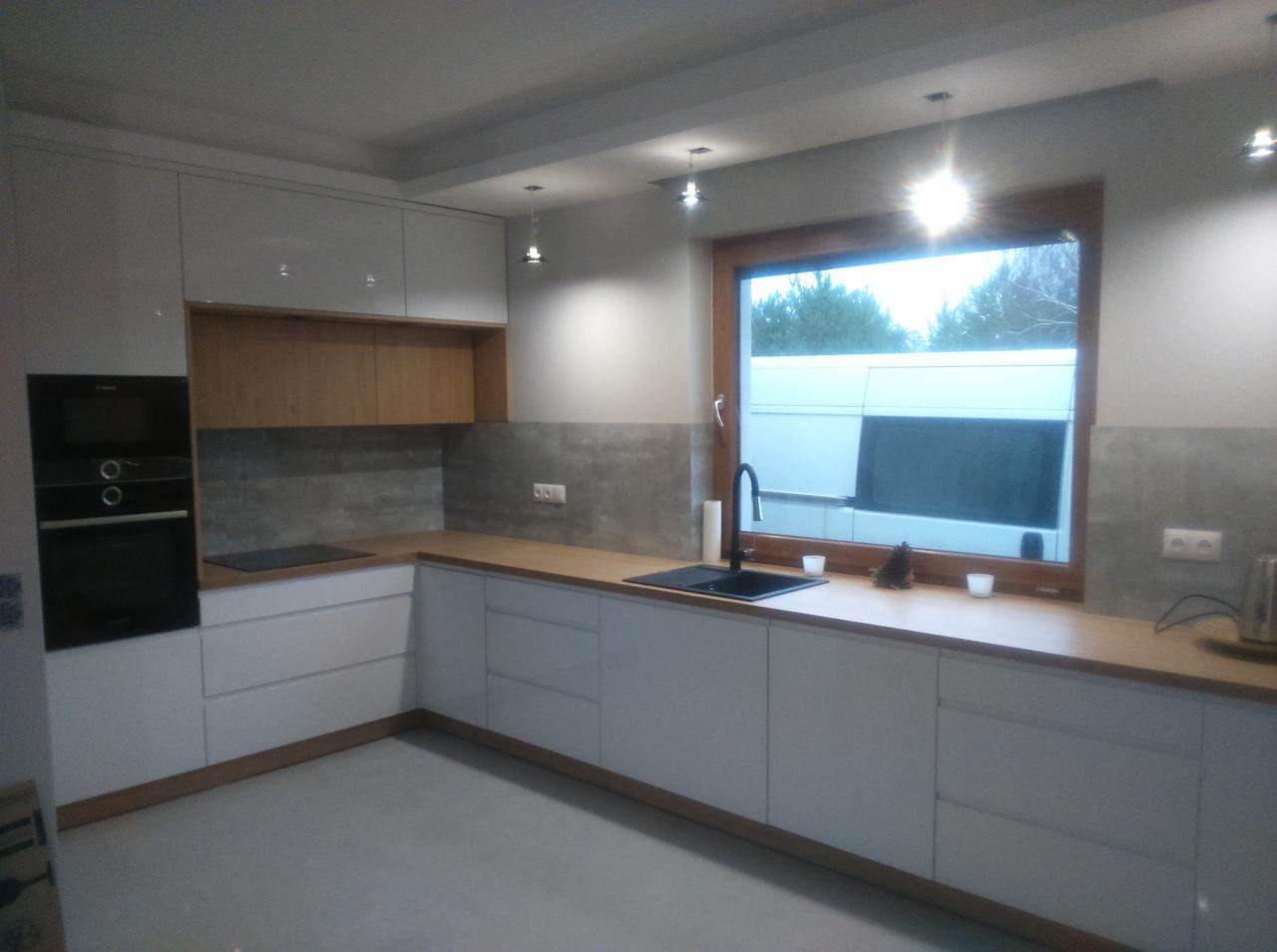 Nowoczesna, jasna kuchnia z białymi, lakierowanymi frontami szafek, drewnianym blatem i szarymi płytkami nad blatem. W tle widoczne okno z czarną armaturą i zabudowa kuchenna z piekarnikiem.