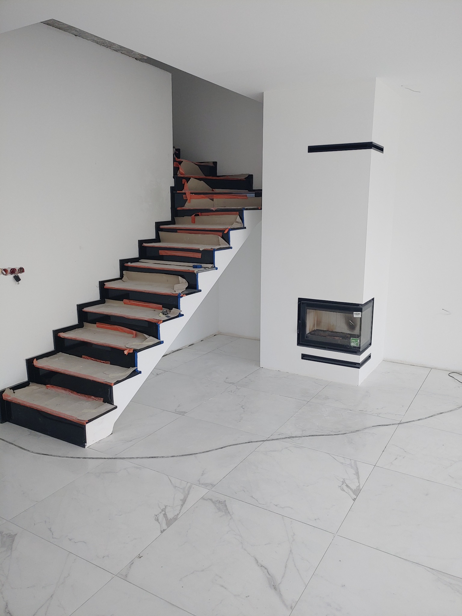 Wnętrze w trakcie prac wykończeniowych: białe ściany, podłoga z płytek imitujących marmur, schody z czarnymi stopniami zabezpieczonymi folią i kominek z czarną obudową.