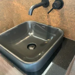 Czarna, prostokątna umywalka ceramiczna Duravit z czarną armaturą Kludi na blacie z czarnego, nakrapianego granitu, zamontowana na szafce z okleiną drewnopodobną, na tle ściany o fakturze miedzi.